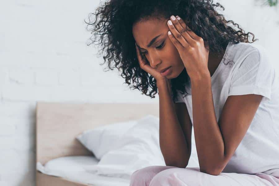Woman-headache-in-pain-holding-head Woman with headache sitting on bed holding her head
